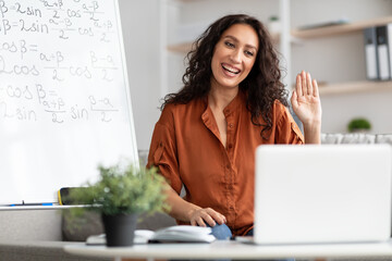 Wall Mural - Smiling young woman teaching maths to students online, waving hello