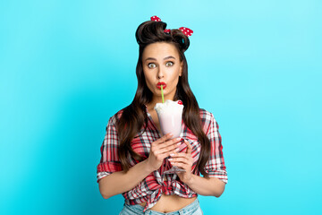 Wall Mural - Shocked young pinup woman in retro clothes drinking yummy milk shake on blue studio background