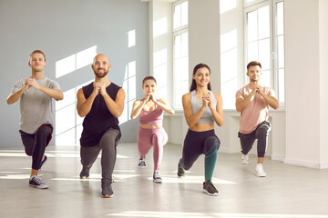 Maintaining good physical condition. Young active people in group sports training together perform leg exercise in bright gym. People going forward lunges holding hands together in front of chest.