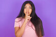 © Jihan - Surprised Hispanic brunette girl wearing pink t-shirt over purple background makes silence gesture, keeps finger over lips and looks mysterious at camera