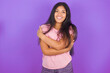 Charming pleased Hispanic brunette girl wearing pink t-shirt over purple background embraces own body, pleasantly feels comfortable poses. Tenderness and self esteem concept