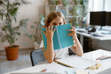 Wall Mural - Shocked little girl hides behind notebook at table in light room