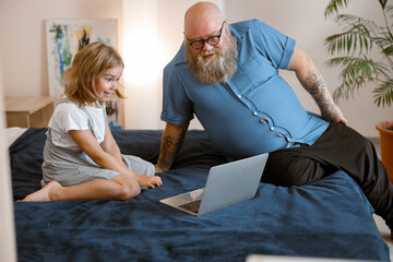 Poster - Bearded father and little girl watch moovie lesson via modern laptop sitting on bed