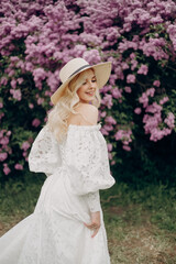 Wall Mural - Young blonde girl in a straw hat and in a white wedding dress with an open top near the lilac bushes