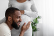 © Prostock-studio - Glad happy millennial african american man listens to belly of pregnant wife in white bedroom interior, close up