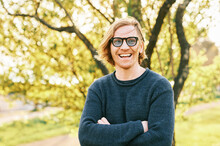 Outdoor Portrait Of Handsome 35 - 40 Year Old Man With Red Hair, Posing In Green Sunny Park, Wearing Blue Pullover And Sunglasses