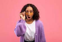 Emotional young African American lady touching sunglasses and looking at camera in shock over pink studio background