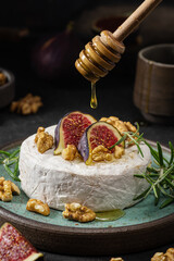Wall Mural - Pouring honey on brie or camembert cheese with figs, walnuts and rosemary on dark background