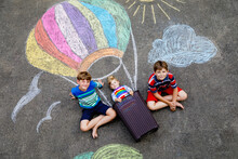 Happy Little Toddler Girl And Two Kid Boys Flying In Hot Air Balloon Painted With Colorful Chalks In Rainbow Colors On Ground Or Asphalt In Summer. Three Children, Siblings Having Fun