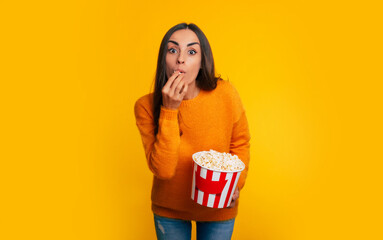 So interesting! Funny young attractive woman in yellow sweater is holding big bucket of popcorn for watching movie in cinema and concentrating looking on camera.