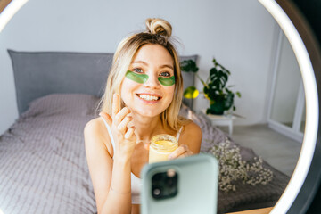 Beautiful woman influencer using her smart phone for a vlog post showing spa facial mask while record and share by smartphone with selfie ring light on social network at home.