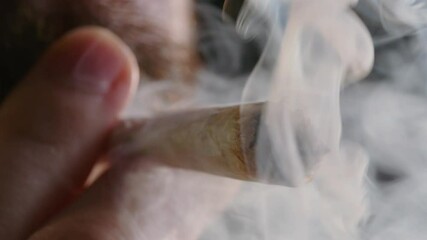 Wall Mural - Extreme close-up shot of male fingers holding lit marijuana joint. Cannabis cigarette burning and a smoke cloud.