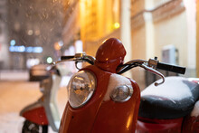 Red Retro Vintage Motor Scooter Under Falling Snow At Night, Selective Focus. Electric Motorcycle Parked In Winter City Lit By Street Lamps. Modern Moped Covered With Snow In Parking Lot In Evening