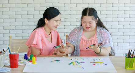 Wall Mural - Asian lovely ponytail hair mother using paintbrush painting acrylic colors on young cute chubby down syndrome autistic daughter hands. Teacher teaching fun art lesson to happy autism artist student