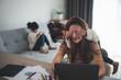 © FAMILY STOCK - woman sits by the table at home office during lockdown, working on laptop. Playful child distracts from work, covering her mother's eyes, kid making noise and asking attention from busy mom
