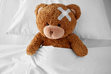 Wall Mural - medicine, healthcare and childhood concept - ill teddy bear toy with medical patch on head lying in bed