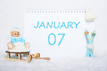 January 7th. Teddy bear sitting on a sled, blue skis and a calendar date on white snow. Day 7 of month.