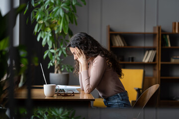 young frustrated female employee getting stuck on task or project, sitting in front of laptop holdin