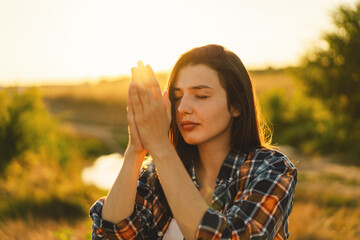 Young woman closed her eyes, praying in a field during beautiful sunset. Hands folded in prayer concept for faith, spirituality and religion. Peace, hope, dreams concept