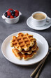 Viennese or Belgian waffles with fresh berries (raspberries and blueberries) on a white plate and a cup of coffee. Traditional dessert. Close-up, grey background.