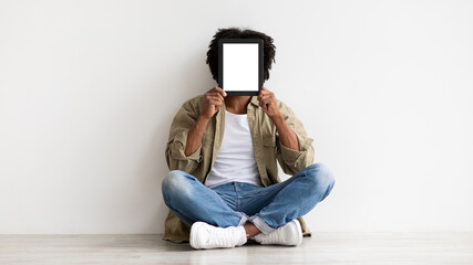 Black Man Covering Face With Blank Digital Tablet While Sitting On Floor