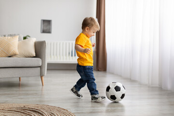 Future champion. Adorable little toddler boy playing football, hitting ball at home, having fun in living room