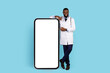 © Prostock-studio - Smiling Black Doctor In Uniform Pointing At Blank Phone With White Screen