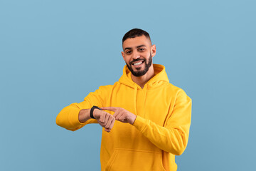 Wall Mural - Happy middle eastern guy touching his smartwatch and smiling at camera, standing over blue studio background