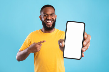 Wall Mural - handsome happy black guy pointing at smartphone with blank screen on blue background, mockup for web