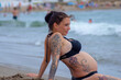 Hübsche dunkelhaarige Frau mit einem Babybauch, ist am rücken und den beinen Tätowiert, liegt am strand und genießt im sommer die wellen.  