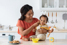 Baby Weaning. Caring Black Mother Feeding Toddler Son From Spoon In Kitchen