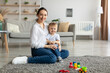 © Prostock-studio - Happy caucasian mother and toddler son smiling at camera, playing together, sitting on floor carpet in living room