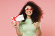 Young curly latin woman 20s wears mint t-shirt sunglasses hold gift certificate coupon voucher card for store show thumb up like gesture isolated on plain pastel light pink background studio portrait.