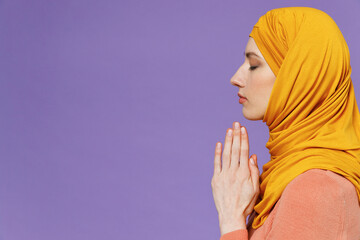 Wall Mural - Side view pleading young arabian asian muslim woman in abaya hijab yellow clothes hands folded in prayer gesture beg about something isolated on plain pastel light violet background studio portrait.