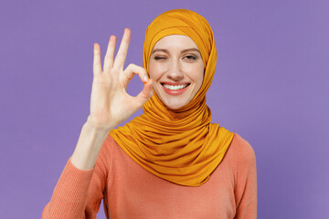 Wall Mural - Smiling happy cheery young arabian asian muslim woman in abaya hijab yellow clothes showing okay ok zero finger gesture wink blink eye isolated on plain pastel light violet background studio portrait.