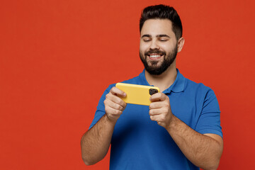 Poster - Young gambling smiling happy caucasian man 20s wear basic blue t-shirt using play racing app on mobile cell phone hold gadget smartphone for pc video games on plain orange background studio portrait