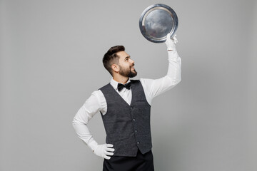 Wall Mural - Young fun barista male waiter butler man 20s wearing white shirt vest elegant uniform work at cafe show hold in hand carrying metal tray isolated on plain grey background. Restaurant employee concept.