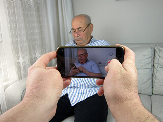   Young youtuber filming a video of an old charismatic man sitting on a sofa and texting on his cell phone. 