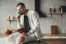 Portrait Of A Man Drinking Coffee Wearing Eyeglasses And Reading A Book While Resting In Kitchen. Coffee Break.