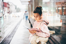 Young Woman Waits For Bus And Reading Notebook On Public Transport Stop Against Large City Blurry Street In Rainy Evening Side View