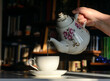 There is a teapot in a woman's hand. She pours tea into a cup.  Background - bookshelves illuminated by the sun.