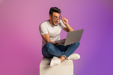 Millennial Arab man sitting cross legged with laptop pc, touching glasses, looking at screen in shock, neon light