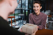 © LIGHTFIELD STUDIOS - Excited woman holding present near wine and blurred boyfriend in hotel cafe