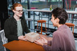 © LIGHTFIELD STUDIOS - Smiling redhead man holding gift near blurred girlfriend and wine in hotel cafe