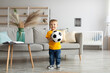 Happy toddler boy football fan posing with soccer ball and smiling at camera, having fun at home in living room