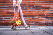 Woman Walking With Reusable Mesh Bag Full Of Fruits. Sustainable Lifestyle, Plastic Free Shopping And Zero Waste Concept. Red Brick Wall Background