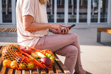 Wall Mural - Woman with reusable mesh bag sitting on bench and using smart phone in city. Resting at bench after shopping fruits in supermarket. Zero waste and plastic free concept