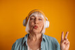 Happy 50s stylish woman giving an air kiss with headphones listening to music and showing peace gesture with hand, enjoying cool playlist