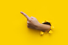 The Forefinger Points To The Left Up Side. Yellow Background. Place For Advertising. Copy Space. The Woman's Hand Came Out Into The Torn Paper Hole.