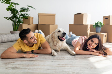 Canvas Print - Cool young diverse couple lying on floor among carton boxes with their cute dog, relocating to new home with pet